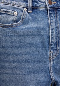 Gros plan sur un jean en denim bleu montrant la poche avant, les passants de ceinture, les rivets métalliques, la zone de la fermeture éclair et les détails de couture orange contrastante.