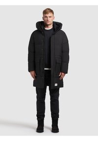 Cappotto invernale imbottito nero con cappuccio, caratterizzato da una zip frontale, ampie tasche frontali e una patch con logo sul orlo. Il design è dritto.
