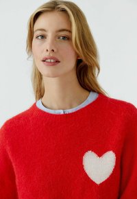 Rood gebreide trui met een wit hartmotief aan de linkerkant. Ronde halslijn met een lichtblauwe kraag en een pluizige textuur.