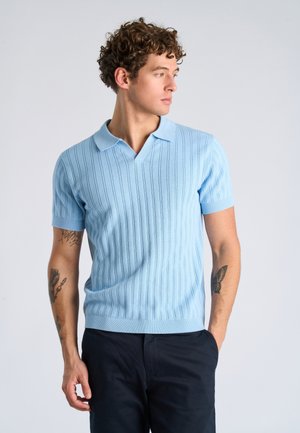 Mężczyzna o kręconych włosach, ubrany w jasnoniebieską, prążkowaną koszulkę polo z krótkim rękawem i ciemne spodnie, patrzący w lewo, z tatuażami na obu ramionach.