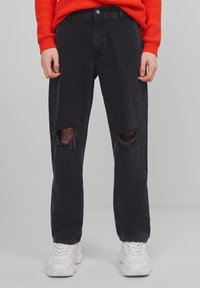 Vaqueros de denim negro con un ajuste relajado, que cuentan con desgarros en las rodillas y un cierre de botón. Llevado con un suéter texturizado rojo y zapatillas blancas.