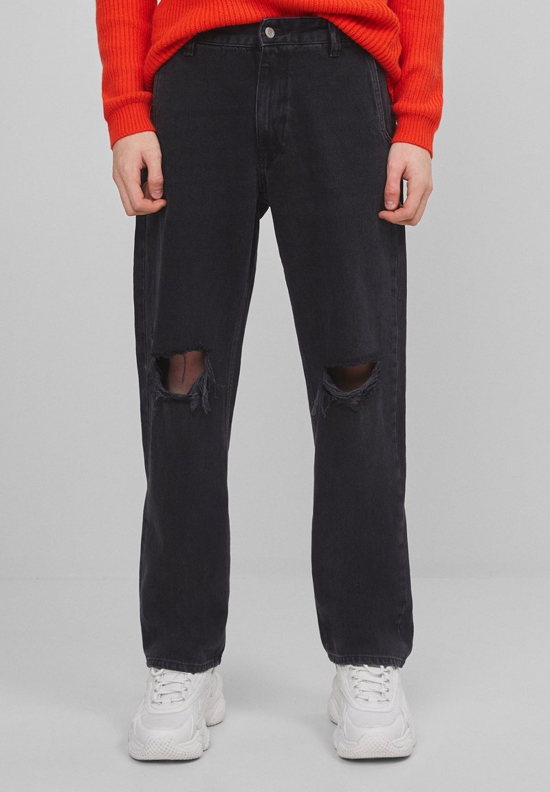 Vaqueros de denim negro con un ajuste relajado, que cuentan con desgarros en las rodillas y un cierre de botón. Llevado con un suéter texturizado rojo y zapatillas blancas.