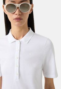 Bílé polo tričko s límečkem, vybavené šestiknutovým zapínáním a malým embosovaným logem. Model nosí kulaté bílé sluneční brýle.