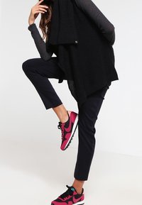 Svart stickad poncho över en mörk topp, parat med marinblå byxor och rosa-svarta träningsskor med en texturerad sula och kontrasterande detaljer.