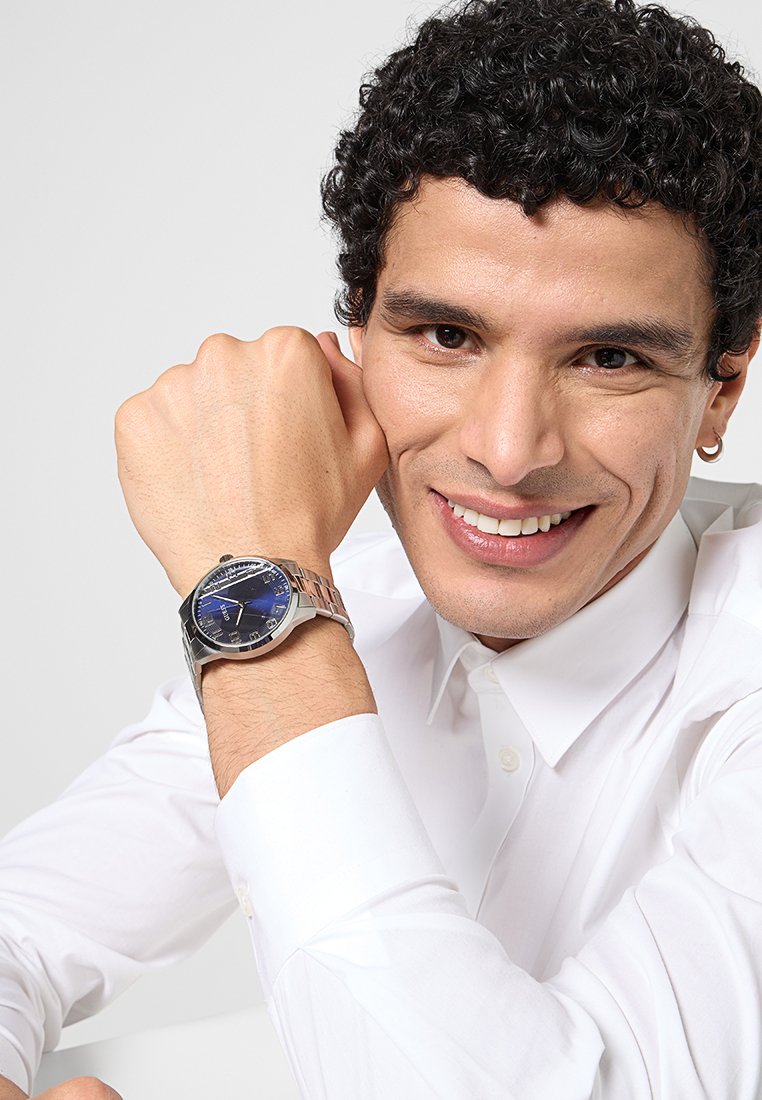 Reloj de acero inoxidable plateado con esfera azul, marcadores de hora plateados y pulsera pulida. El portador posa con la mano apoyada en la barbilla.