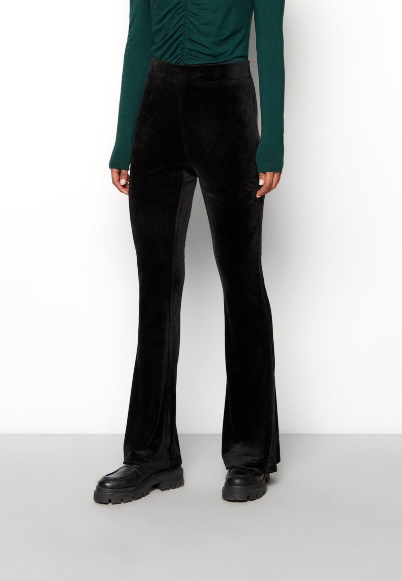 Personne portant un pantalon droit en velours noir, des chaussures noires épaisses et un haut froncé à manches longues vert foncé, debout sur un sol gris.