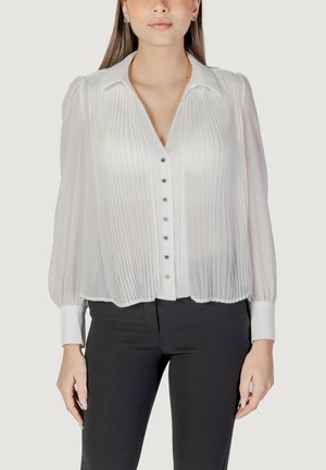 Blusa bianca con pieghe, maniche lunghe e colletto, caratterizzata da una chiusura frontale con bottoni e bottoni argentati, abbinata a pantaloni scuri.