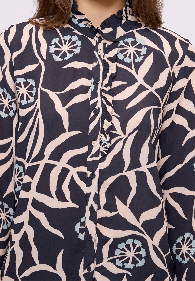 Blouse noire avec motif floral abstrait beige et bleu clair, avec col froufroutant et fermeture boutonnée devant.