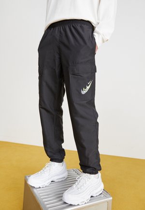 Person iført sorte cargobukser med flere Nike-logoer på lommen og hvide sneakers, stående på et metaltrin mod en ensfarvet baggrund.