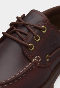 Κοντινό πλάνο καφέ δερμάτινου παπουτσιού για βάρκα με ορειχάλκινες τρύπες, καφέ κορδόνια, λευκή ραφή και ανάγλυφο λογότυπο Timberland στο πλάι.