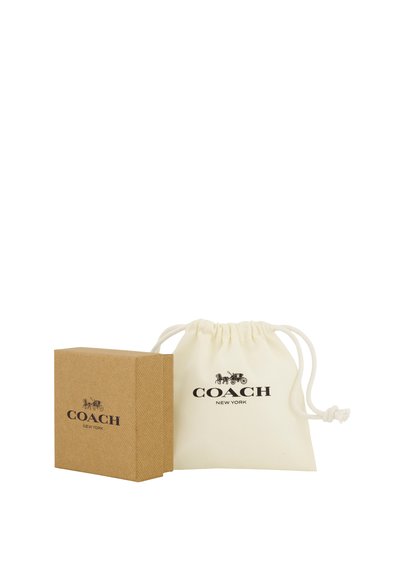 Une boîte Coach New York couleur fauve est posée à côté d’une pochette crème à cordon de serrage. Les deux affichent le logo de la marque sur un fond blanc.