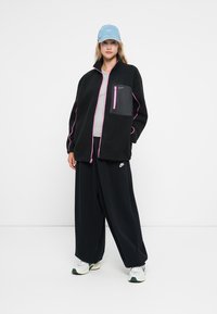 Crna flis jakna s ružičastim detaljima i zatvorenim crnim džepom. Uparena s rebrastim crnim hlačama i svijetloplavom kapom s Nike logom.