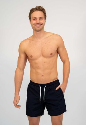 Lachende jonge man met kort blond haar, zonder hemd, gekleed in zwarte zwembroek met witte trekkoorden, staand tegen een effen lichte achtergrond.