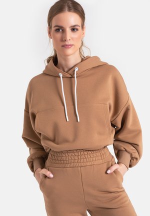 Brązowa bluza z kapturem o luźnym fasonie, z białymi sznurkami i ściągaczami na rękawach. Posiada kieszeń kangurkową oraz miękki, przypominający polar materiał.