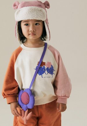 Ein Kind trägt einen mehrfarbigen Sweatshirt mit blauen Cartoonfiguren und dem Text "BEST FRIEND". Kombiniert mit orangefarbenen Hosen und einer lila Blumenhandtasche.