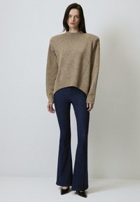 Maglione beige lavorato a maglia con collo alto, spalle cadenti e orlo curvo, abbinato a jeans a vita alta scuri e tacchi a punta neri.