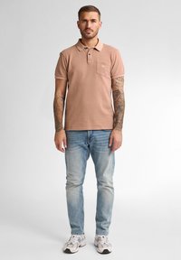 Béžové polo tričko s texturovanými pruhy, krátkými rukávy a kapsou na prsou. Nosené s světle modrými džíny a bílými teniskami.