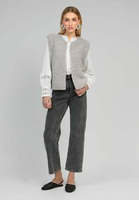 Gilet en tricot gris sur une chemise blanche à boutons avec des détails en dentelle, associé à un jean noir taille haute et des chaussures noires à talons ouverts.
