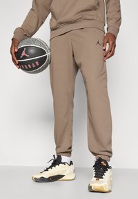 Brune joggebukser med elastiske mansjetter, med en liten svart Jordan-logo. Personen holder en teksturert basketball med røde detaljer.