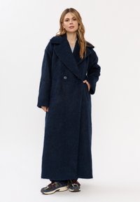 Donna che indossa un lungo cappotto peloso blu navy con ampie rever, pantaloni scuri e sneaker multicolori, in piedi su uno sfondo bianco.