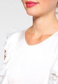 Blusa bianca con dettagli arricciati sulle spalle, texture liscia e scollatura rotonda, che mette in evidenza un design semplice ma elegante.