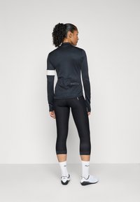 Zwart sporttop met lange mouwen en een witte accent op de linkerarm, gecombineerd met zwarte aansluitende leggings en witte sportschoenen.