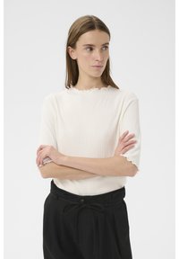 Biele rebrované tričko s jemným volánovým výstrihom a krátkymi rukávmi, nosené k vysokým čiernym nohaviciam s viazaním v páse.