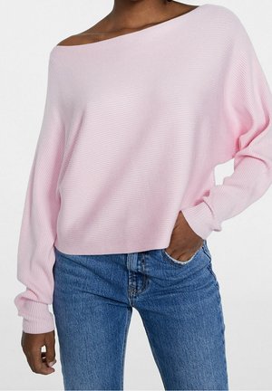 Vrouw draagt lichtroze off-shoulder geribbelde trui met lange mouwen en blauwe spijkerbroek, één hand in de zak, tegen een effen achtergrond.