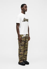 Λευκό μπλουζάκι με μαύρο και πολύχρωμο γραφικό σχέδιο, σε συνδυασμό με παντελόνι cargo στο μοτίβο παραλλαγής σε πράσινο χρώμα και σκούρα υποδήματα με σχέδιο.