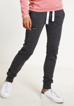 Person iført mørkegrå joggebukser med hvide snore, en lyserød sweatshirt og hvide sneakers med sorte mønstrede detaljer.