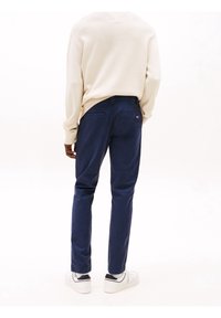 Persona con un suéter crema, pantalones azul marino y zapatos deportivos blancos de pie de espaldas contra un fondo blanco.