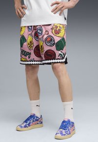Pantalones cortos de baloncesto rosas con varios gráficos coloridos, incluidos un gato, balones de baloncesto y latas verdes. Detalle de dobladillo en zigzag negro. Llevados con zapatillas de deporte de diseño.