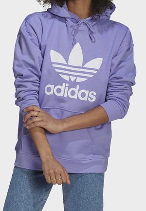 Personne portant un sweat à capuche Adidas violet avec logo blanc, jean bleu clair et plusieurs bagues aux doigts, bras croisés à la taille.