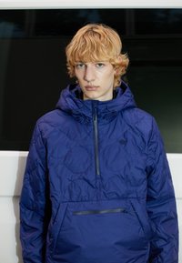 Giubbotto puffo viola con cappuccio, dotato di tasca frontale con zip e una superficie liscia e lucida. Presenta un piccolo logo sul petto.