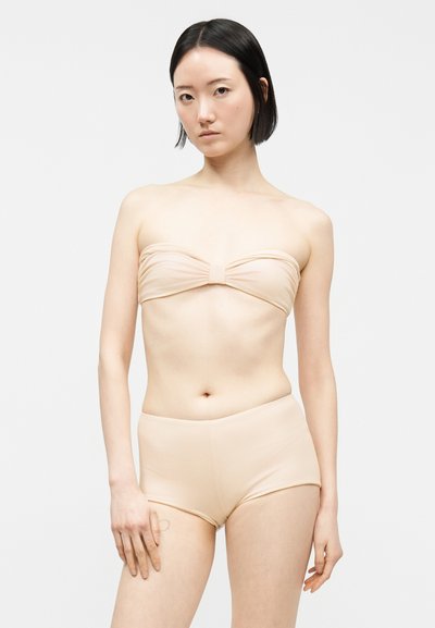 Νεαρή γυναίκα με κοντά μαύρα μαλλιά, που φοράει μπεζ κορμάκι χωρίς τιράντες και σορτς με ψηλή μέση, σε λευκό φόντο.