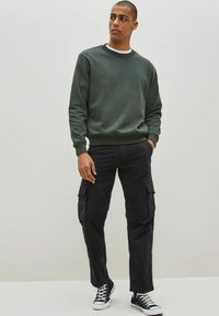 Ciemnozielona bawełniana bluza z okrągłym dekoltem, zestawiona z czarnymi spodniami cargo z bocznymi kieszeniami, noszona z czarnymi sneakersami za kostkę.