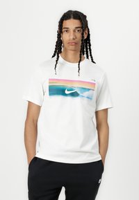 Biely bavlnený tričko s farebnou grafickou potlačou, ktorá zobrazuje oceánske vlny a logo Nike. Krátke rukávy a okrúhly výstrih.
