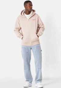 Sudadera beige con cremallera y capucha ajustable, bolsillos tipo canguro y logo bordado. Combinada con pantalones cargo de color azul claro y zapatillas blancas.