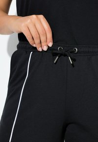 Czarne bawełniane joggery z regulowanym pasem na sznurek ozdobionym metalowymi elementami oraz białym lampasem po bokach na całej długości. Zbliżenie na rękę regulującą pas.