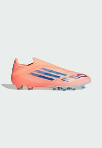 Bota de fútbol de coral brillante con rayas azules, diseño sin cordones y tacos translúcidos sobre un fondo claro y liso.