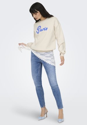 Vrouw draagt beige sweater met opschrift "Paris", blauwe spijkerbroek en lichtblauwe hakken, houdt witte kanten stof diagonaal over haar heupen.