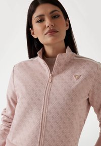 Veste zippée rose avec un motif en losanges, des poignets côtelés et un petit logo sur la poitrine. Fabriquée dans un matériau doux avec un col montant.