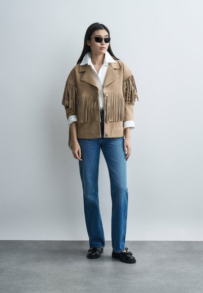 Vrouw met zwarte zonnebril, een tan kleurige jas met franjes, wit shirt, blauwe jeans en zwarte schoenen, staand tegen een effen lichtgrijze achtergrond.