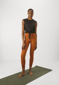 Fekete ujjatlan felső, csomózott részletekkel, narancssárga leggings-szel párosítva, amelyen egy diszkrét logó látható. Zöld jógaszőnyegen áll, mezítláb.