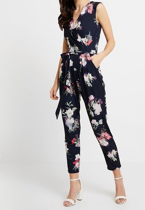 Femme portant une combinaison sans manches bleu marine à imprimé floral rose et blanc, taille ceinturée, et des talons blancs ouverts, posant devant un fond uni.