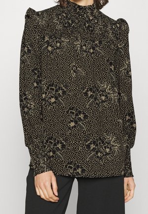 Woman wearing black long-sleeve blouse with beige floral and dotted patterns, high ruffled collar, and puffed shoulders.
