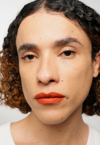 Nærbilde portrett med krøllete brunt hår, lys hud, definerte trekk, og matt oransje leppestift. Subtil hudtekstur og naturlig sminke.