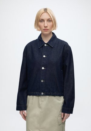 Woman with short blonde hair wearing a dark blue buttoned denim jacket and a light beige skirt, facing forward with a neutral expression.