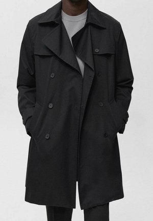 Homme portant un trench-coat noir croisé avec les mains dans les poches sur un pull gris clair.