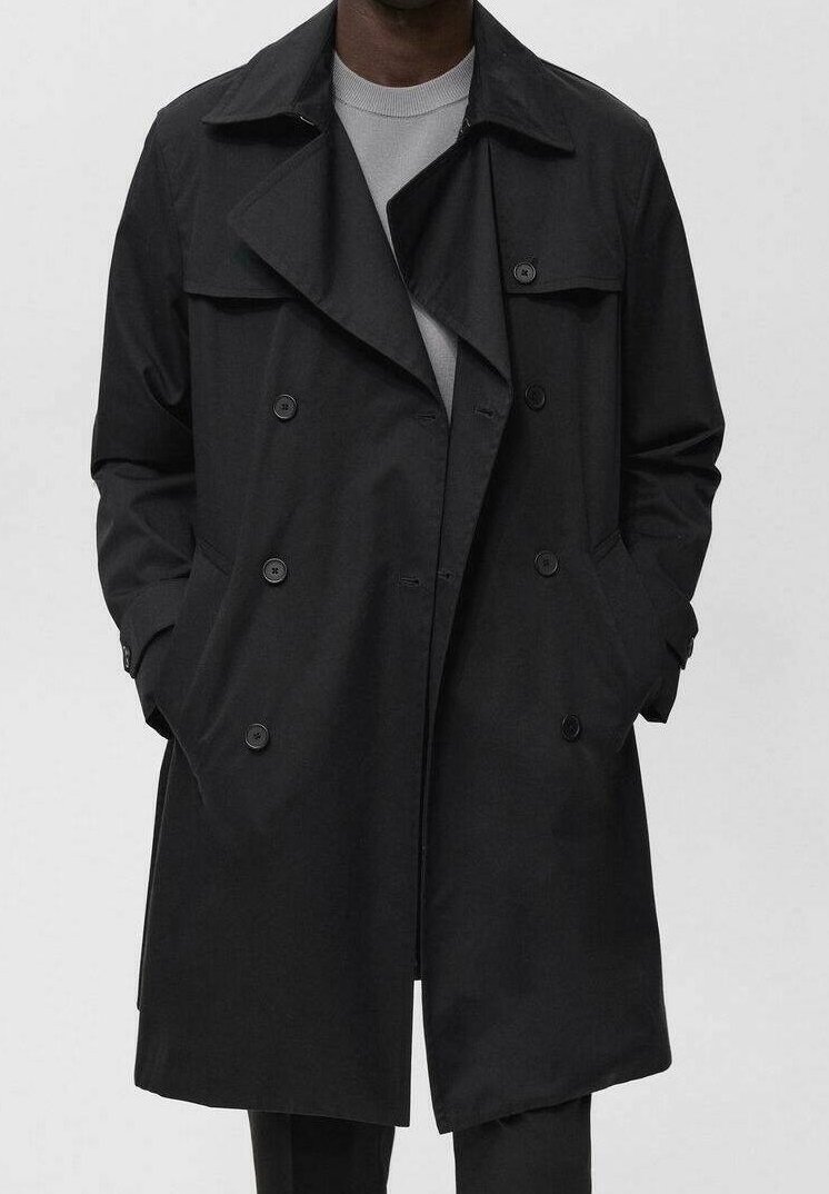 Homme portant un trench-coat noir croisé avec les mains dans les poches sur un pull gris clair.